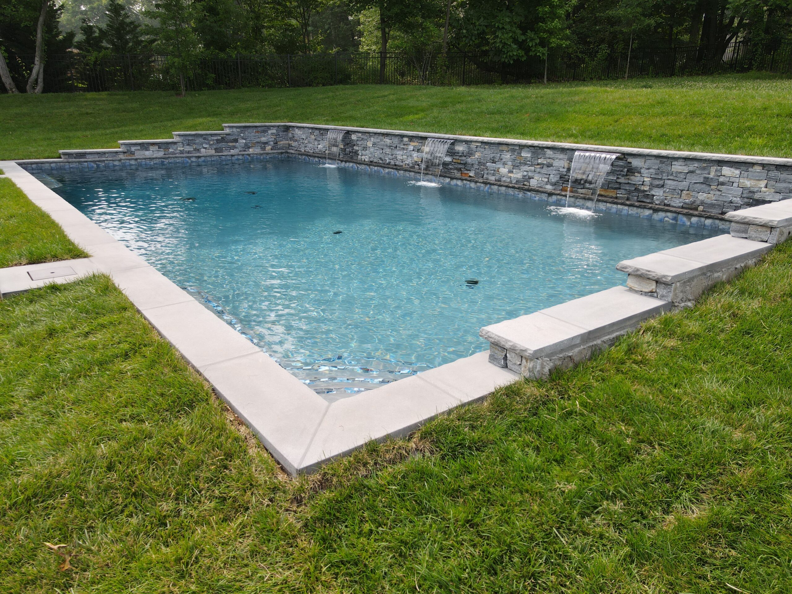 A backyard rectangular swimming pool with a stone water feature and a tiered rock wall. The pool has a light blue interior and is surrounded by a well-manicured lawn and a light gray paver deck. The coping is also a light gray stone. The stone wall features three rectangular water features from which water spills into the pool.