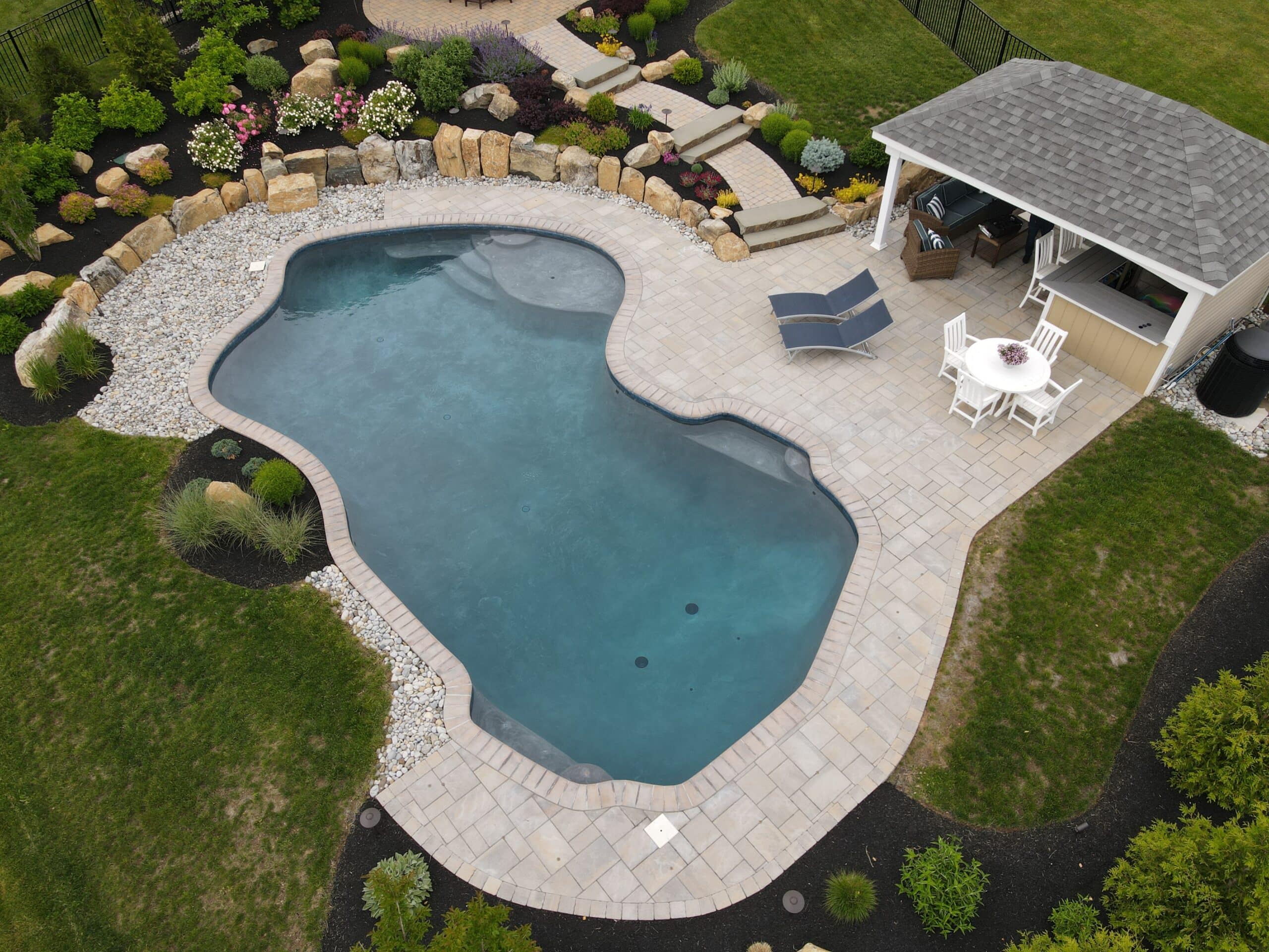 An aerial view of a backyard with a freeform swimming pool and a small, attached pergola. The pool has a dark blue interior and is surrounded by a light-colored paver deck. A set of stepping-stone stairs leads up a small, landscaped hill with a rock garden to a grassy area.