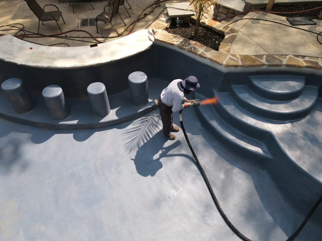 A high-angle shot of a man wearing a hat and face mask, working on an empty, freeform swimming pool. He is using a tool that emits a flame to coat the pool's surface near a set of built-in steps. Several gray cylindrical stools are visible on a ledge on the left side of the pool. The pool deck is a mix of concrete and stone pavers, with a few chairs and a small palm tree visible in the background.