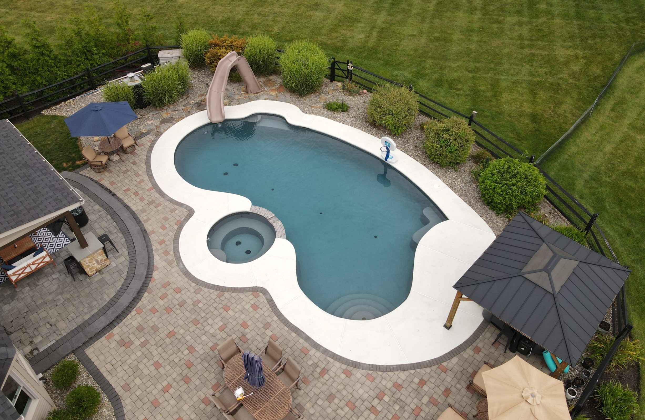 An overhead view of a large, custom-shaped backyard swimming pool with a connecting hot tub. The surrounding patio is a light concrete. The pool area features a hot tub, a built-in slide, a basketball hoop, and several seating areas, including a covered gazebo and a patio dining set.