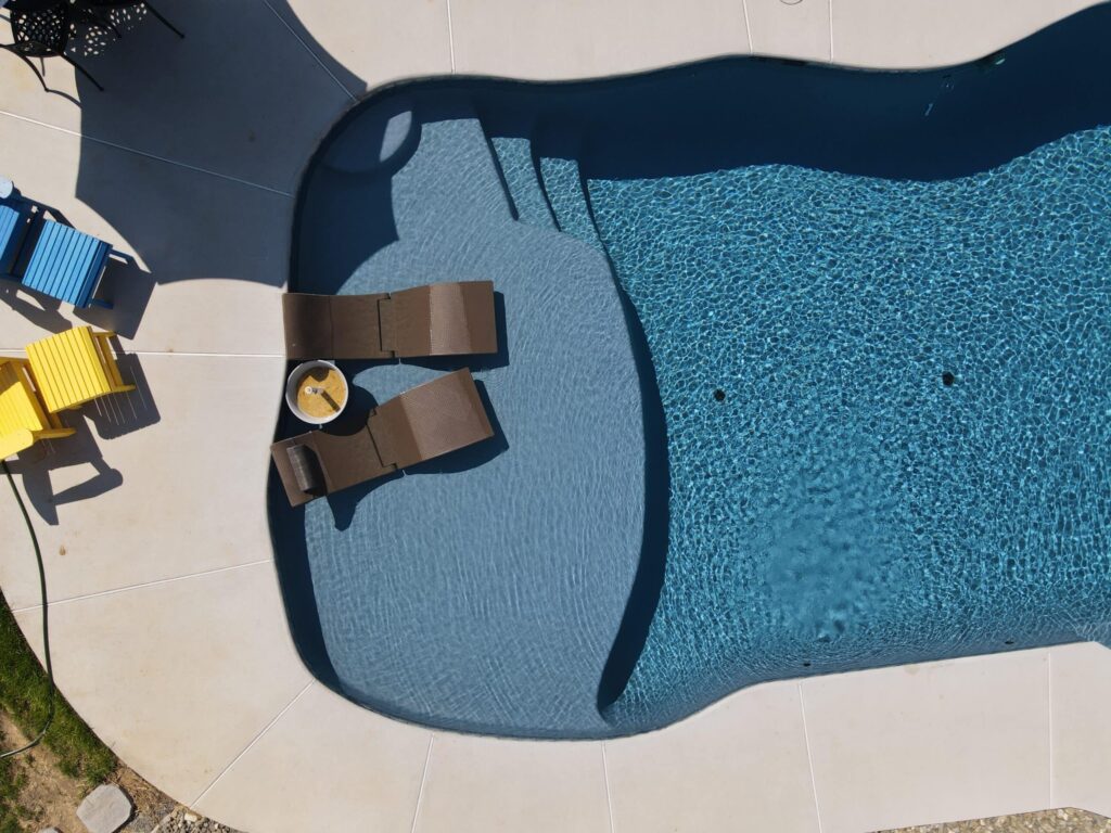An aerial view of a freeform swimming pool with a unique shallow end that has a built-in sun shelf. The sun shelf, with its smooth, light gray surface, has two brown lounge chairs on it and a small umbrella stand. The main pool area is a darker, textured blue and has steps leading into it. On the light-colored concrete deck, there are several colorful Adirondack-style chairs in blue and yellow.