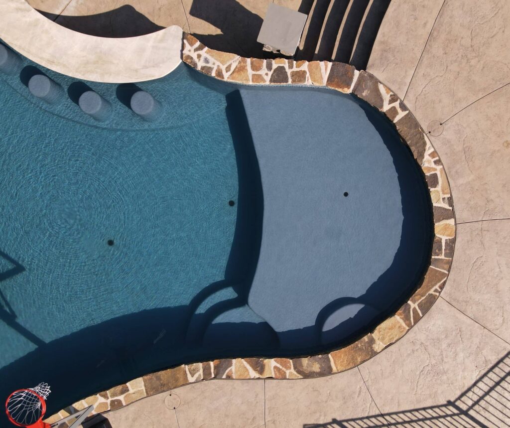 An overhead shot of a swimming pool sun shelf. The pool has a dark blue liner, and its edges are lined with a decorative, multi-toned stone coping. The surrounding deck is a light brown, stamped concrete. In the shallow end of the pool, there are built-in stools, and a small, raised platform with a lighter-colored stone border.