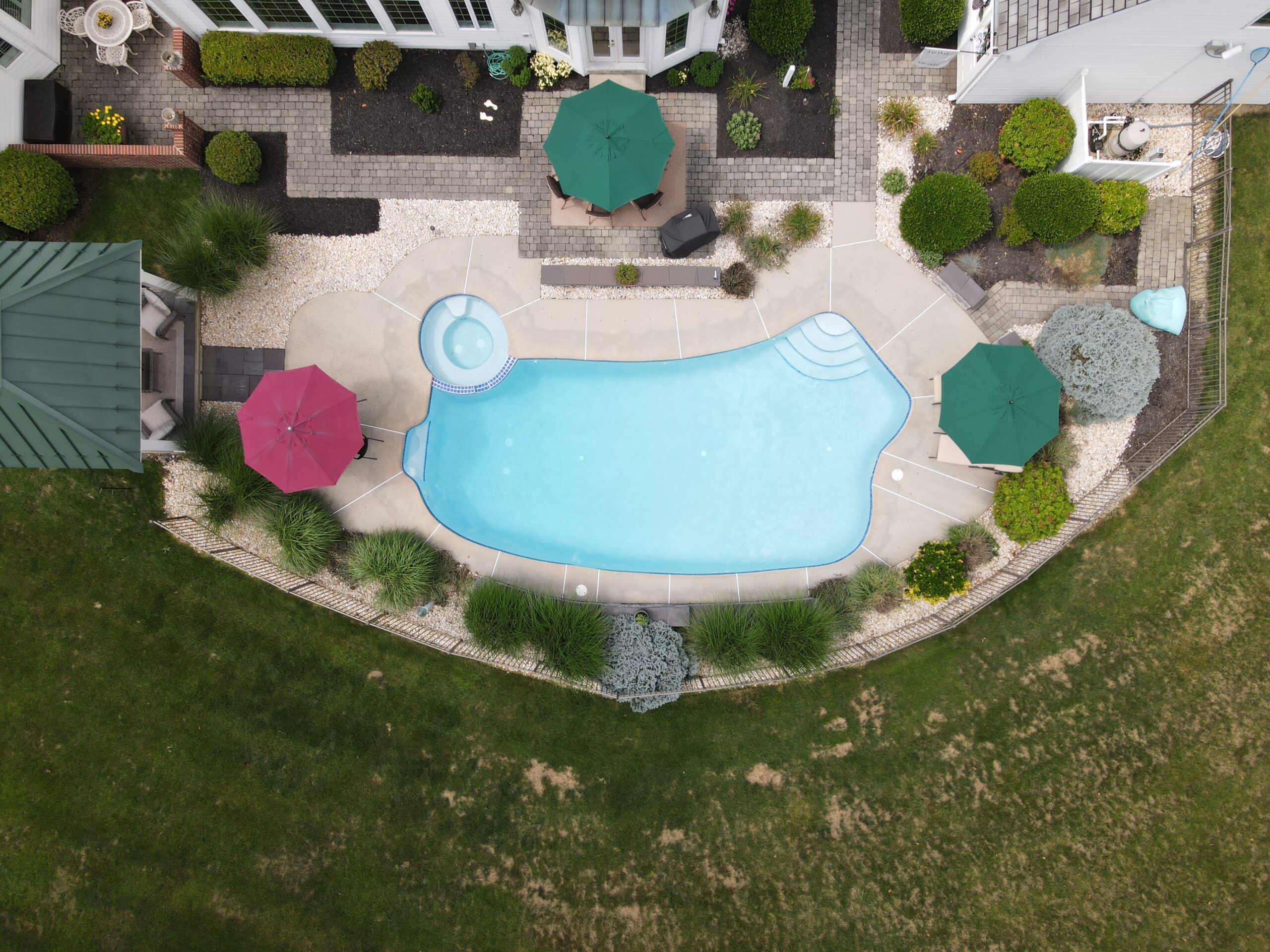 An aerial shot of a large backyard with a bean-shaped swimming pool. The pool has a curved set of steps and is surrounded by a light-colored concrete deck. The deck has a connected circular hot tub area.