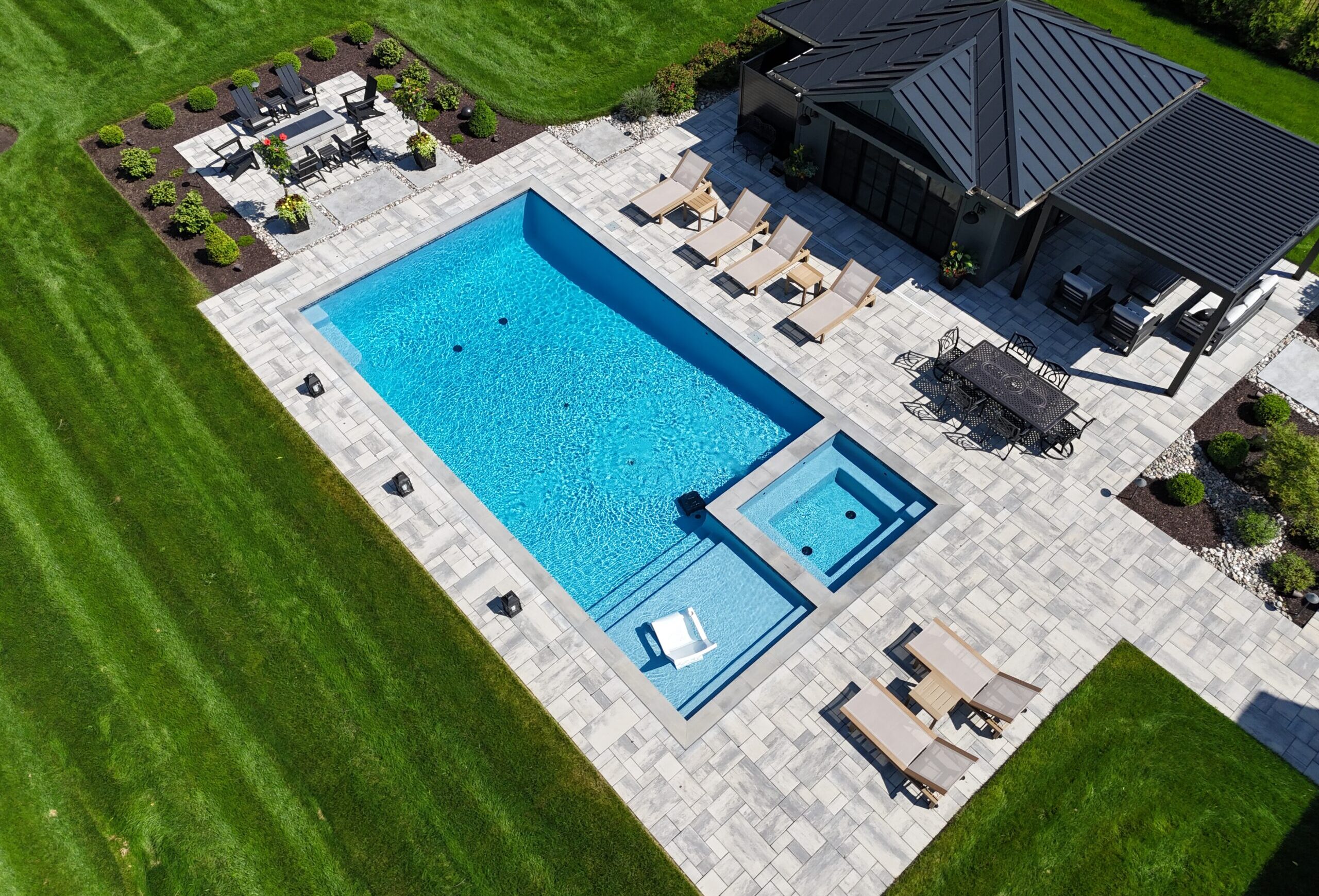 An elevated, overhead view of a modern backyard pool area. The pool is a rectangular shape with a light blue interior. A square spa is attached to the main pool. The entire area is surrounded by a light-colored paver patio, with a dining set and several lounge chairs. The yard is a mix of green grass and landscaped garden beds.