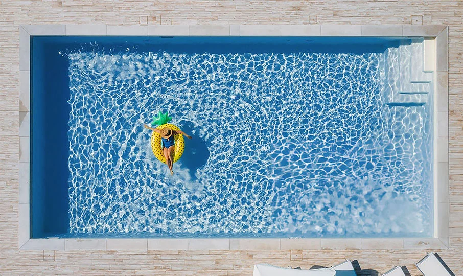An aerial shot of a woman floating on a large, inflatable yellow pineapple raft in a rectangular swimming pool. The pool has a deep blue interior and is surrounded by a light-colored, tiled patio. Steps are visible in the corner of the pool.