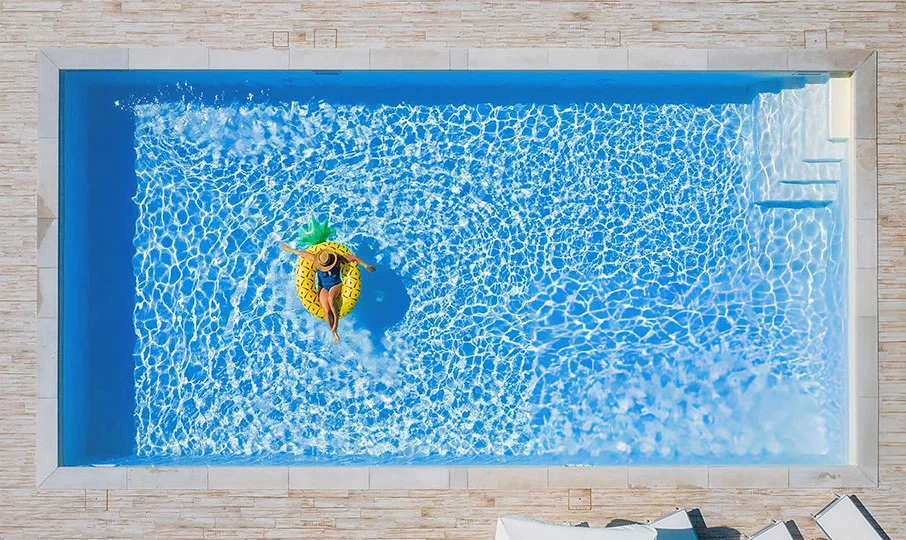 An aerial shot of a woman floating on a large, inflatable yellow pineapple raft in a rectangular swimming pool. The pool has an ocean blue interior and is surrounded by a light-colored, tiled patio. Steps are visible in the corner of the pool.