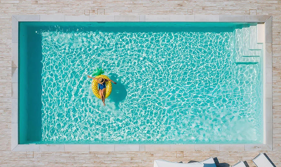 An aerial shot of a woman floating on a large, inflatable yellow pineapple raft in a rectangular swimming pool. The pool has a teal interior and is surrounded by a light-colored, tiled patio. Steps are visible in the corner of the pool.
